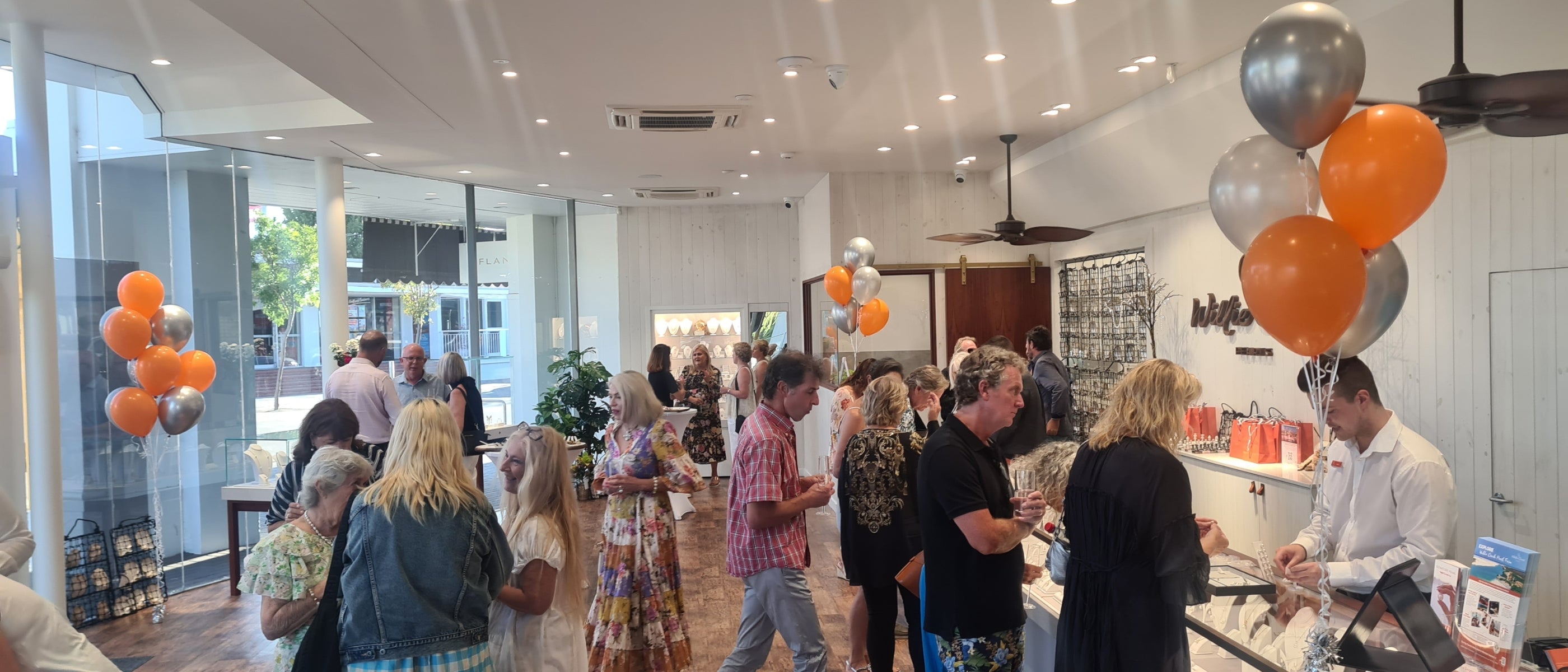 Colourful silver and orange balloons decorate the Cottesloe Willie Creek Pearls showroom, where guests mingle, hold drinks, and shop for pearl jewellery during an event.