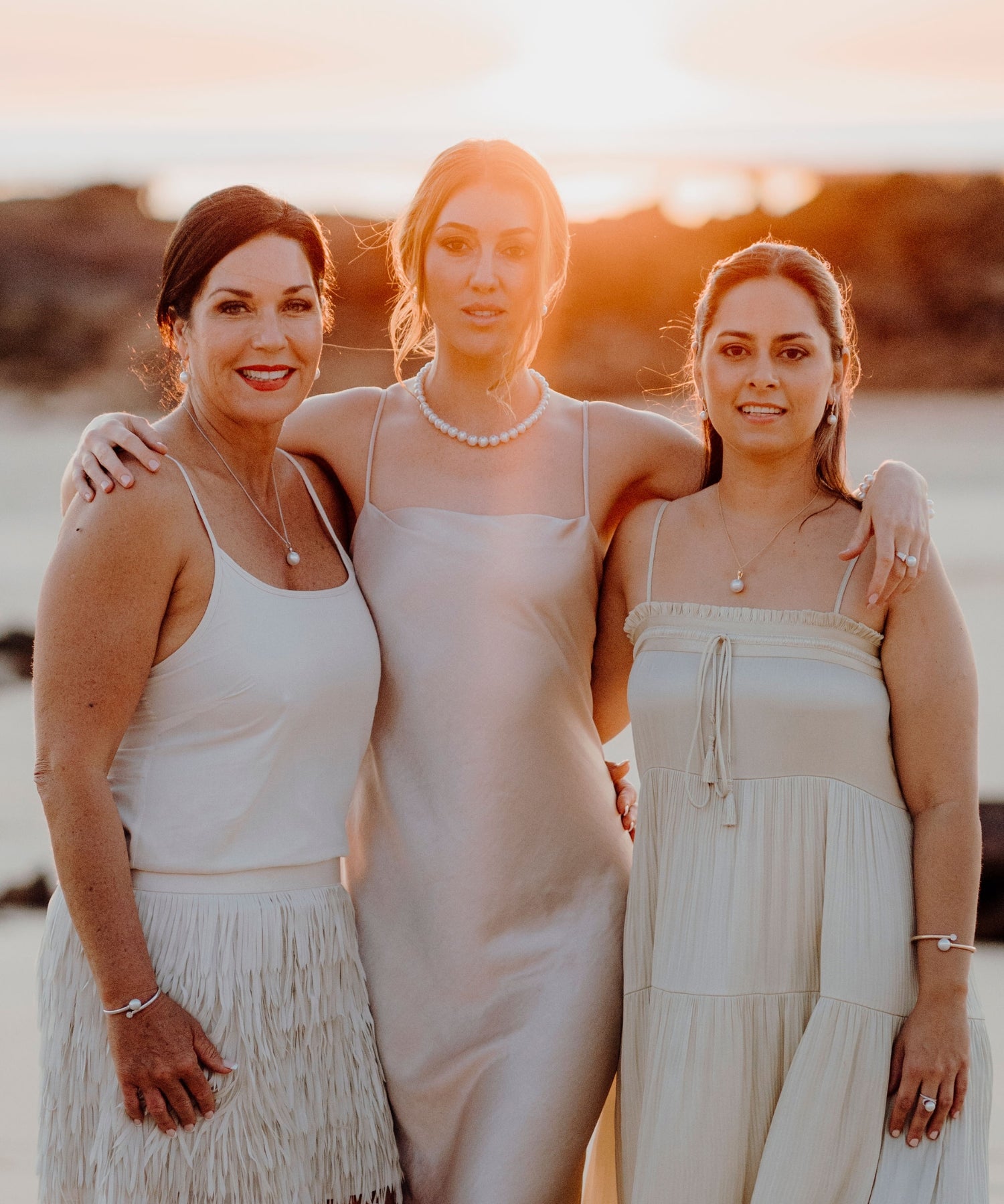 Three models stand in front of the ocean in Broome, wearing elegant cocktail dresses and the Willie Creek Pearls Event Pearl Jewellery Collection, showcasing pearl pieces perfect for special occasions.