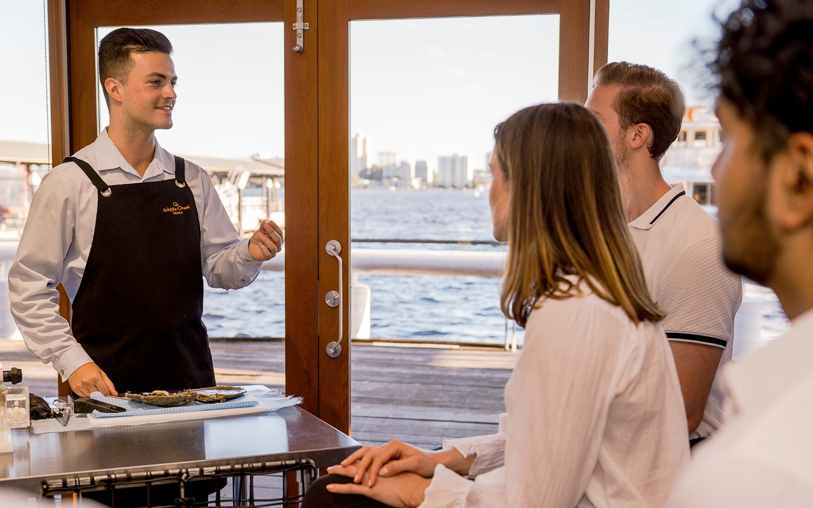 A tour guide stands before a Perth river view, holding a pearl from a freshly harvested oyster, showing it to a small audience as part of the Harvest Your Own Pearl experience.