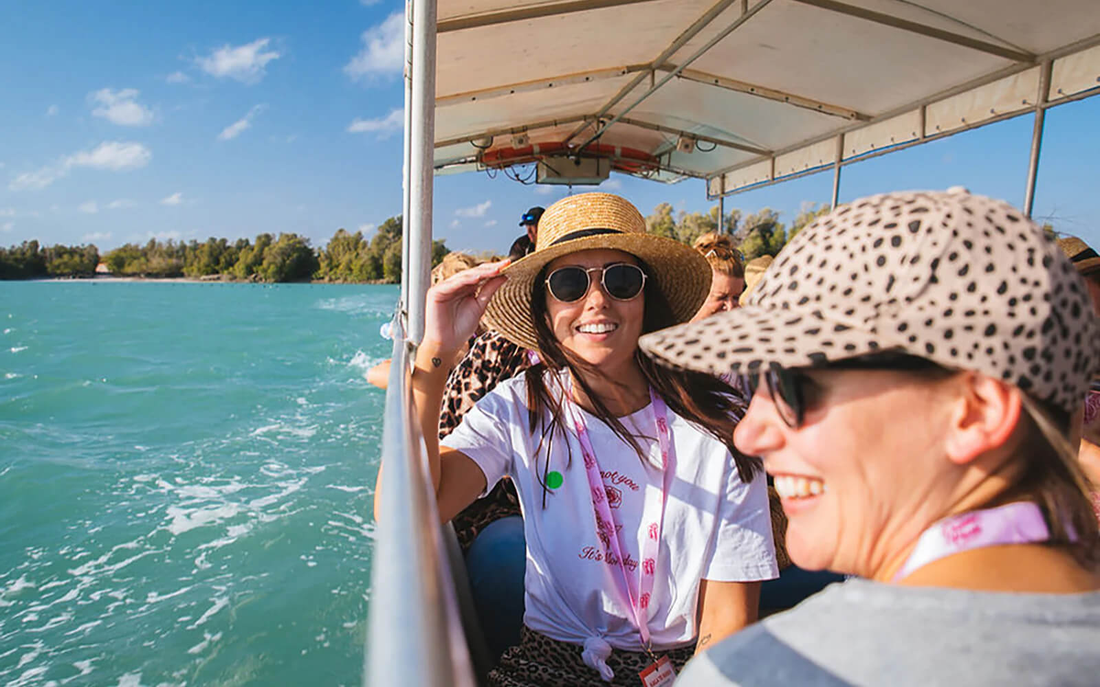 Two women wearing hats and sunglasses smile while enjoying a boat cruise on Willie Creek in Broome, Western Australia, with scenic water views in the background.