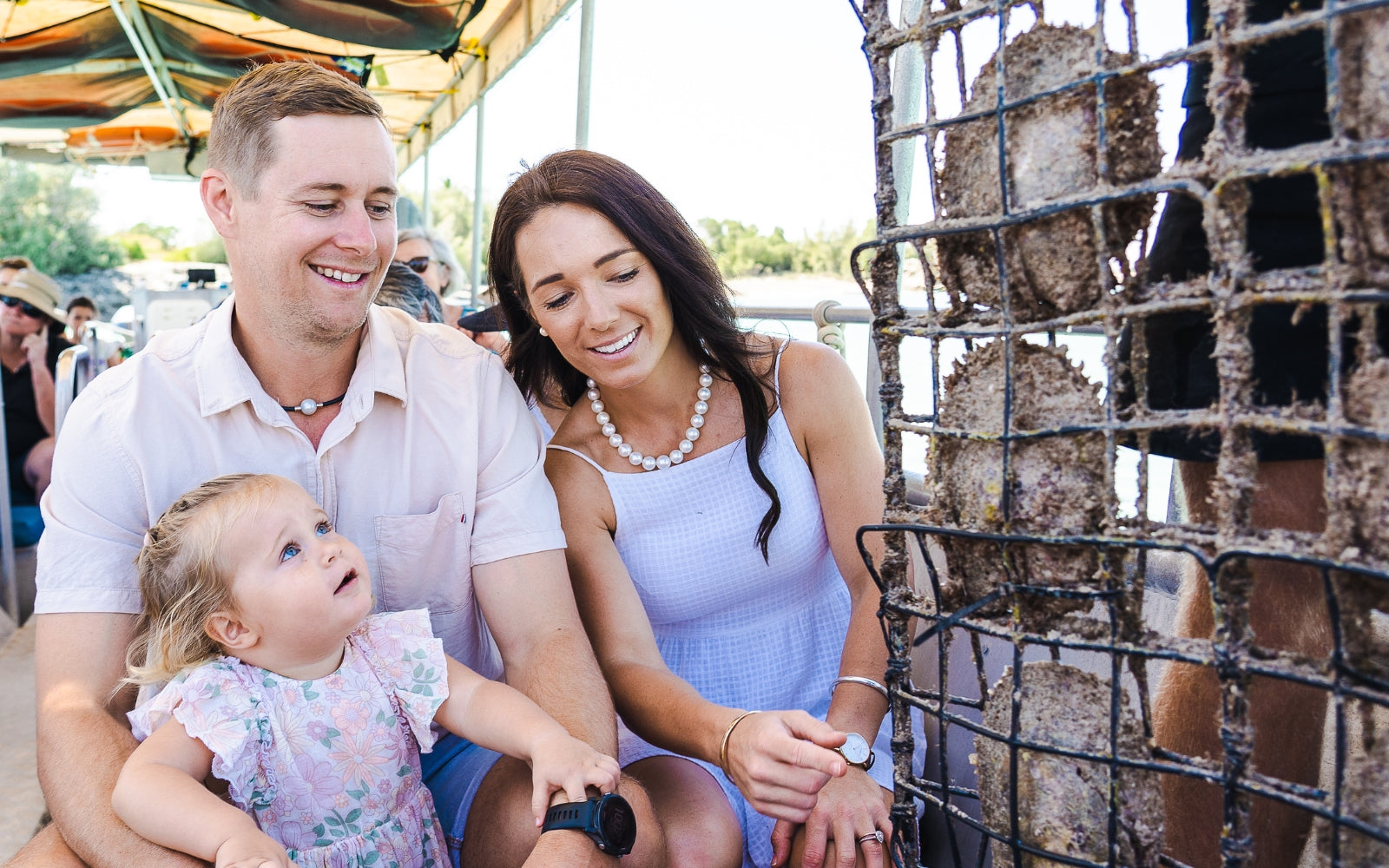 A family wearing pearl jewellery smiles on the Willie Creek Pearl Farm tour boat next to a panel of oysters, observing the pearl cultivation process.