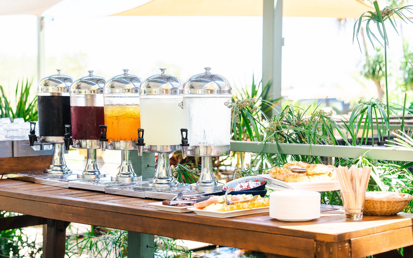 Morning tea setup on the Willie Creek Pearl Farm tour with colourful drink dispensers, damper, fresh fruit, and utensils on a wooden table in an outdoor setting.