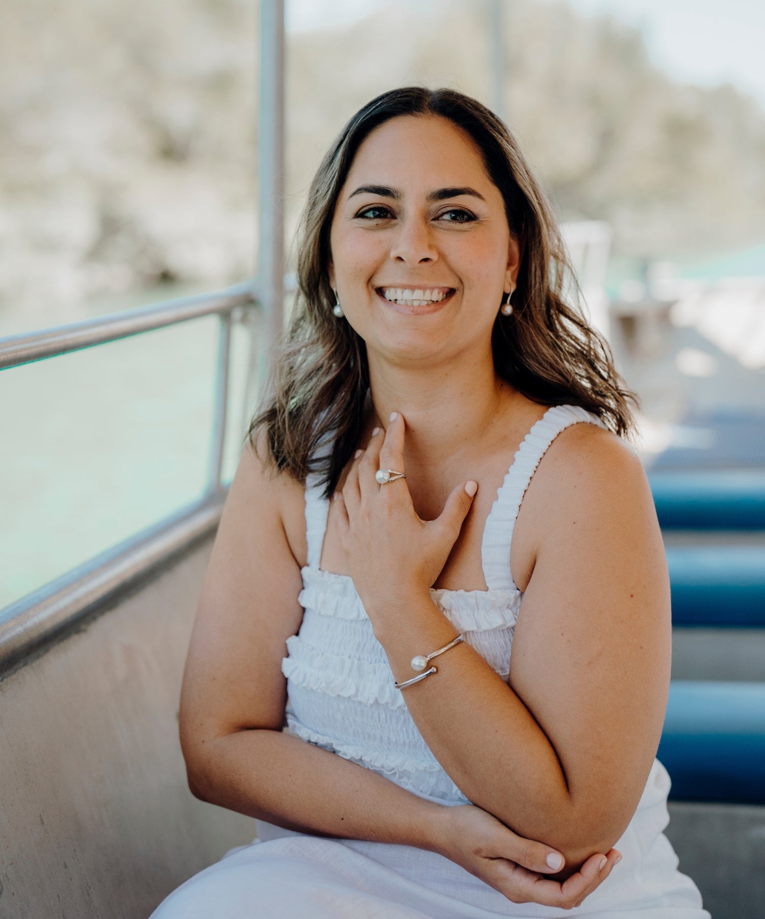 A smiling woman wearing a white dress sits on a boat, showcasing pieces from the Willie Creek Pearls Gift Range, including a pearl ring, bracelet, and earrings.