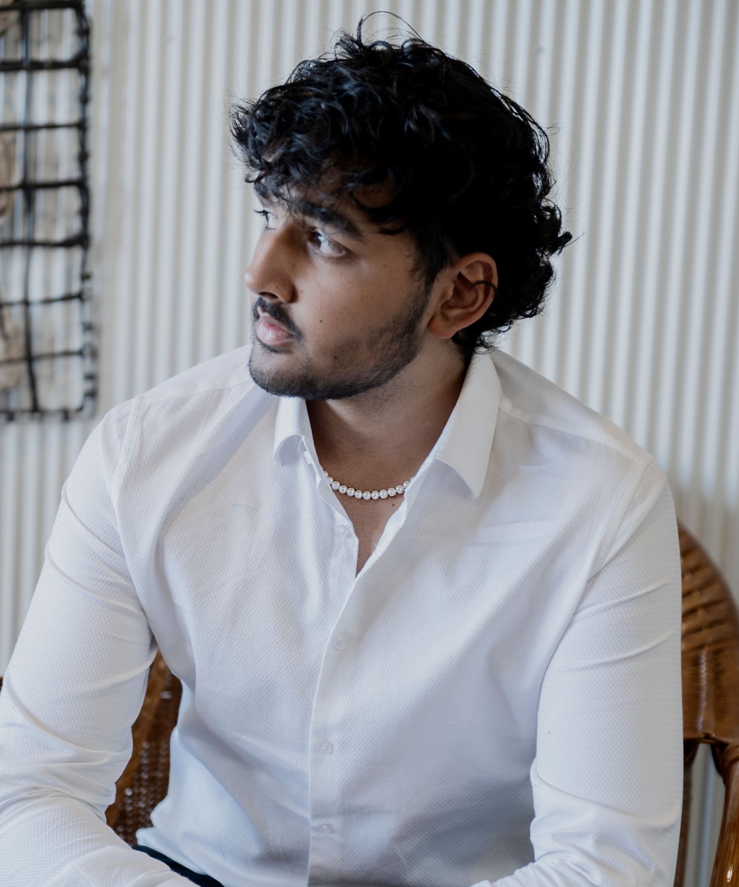 A man wearing a white shirt sits in a relaxed pose, wearing a pearl necklace from the Willie Creek Pearls Men's Collection.