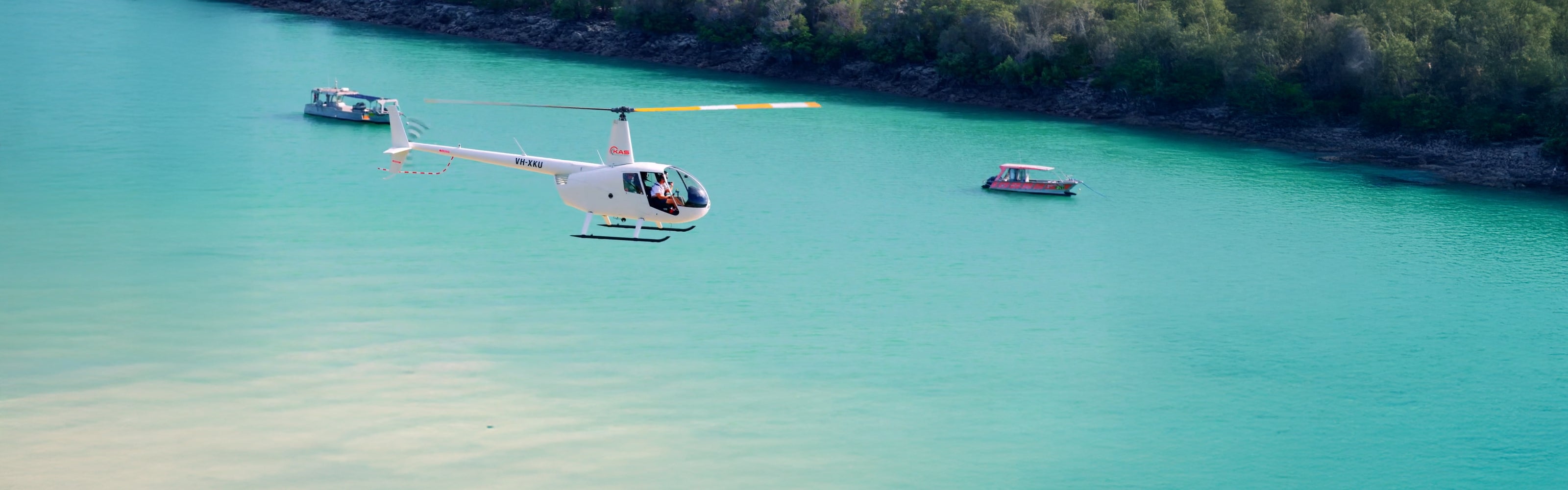 A white helicopter hovers over two boats in the turquoise blue waters of Willie Creek by the Willie Creek Pearl Farm in Broome